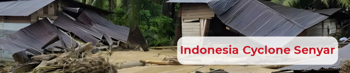 Several wooden houses with corrugated metal roofs collapsed and partially buried in mud and debris. Large tree trunks and branches are scattered across the scene, from Cyclone Senyar in Indonesia. Dense green vegetation is visible in the background.