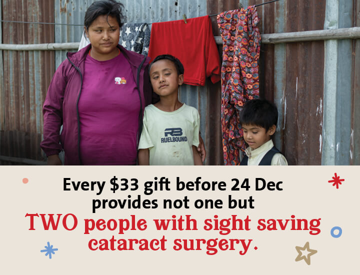 A woman in a purple top stands with three children in front of a corrugated metal wall, with colorful clothes hanging behind them. Text below reads: 'Every $33 gift before 24 Dec provides not one but TWO people with sight saving cataract surgery'
